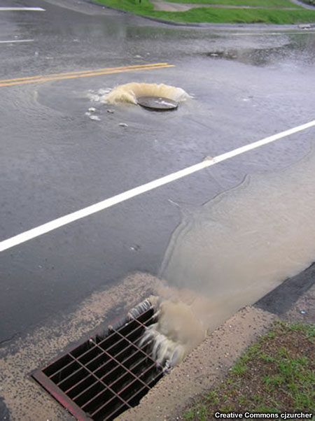 Rainwater flowing down a street and through a storm drainage grate.  Nearby, water is flowing up and out of a manhole cover and onto the street again